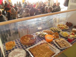 Im Vordergrund ein Buffet mit Kuchen und Käsefüßen, im Hintergrund sitzen Zuhörer.