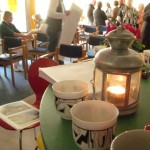 Foto von Kaffeetassen, Kerzen, Primeln im Vordergrund auf dem Tisch, im Hintergrund in Gruppen diskutierende Menschen.