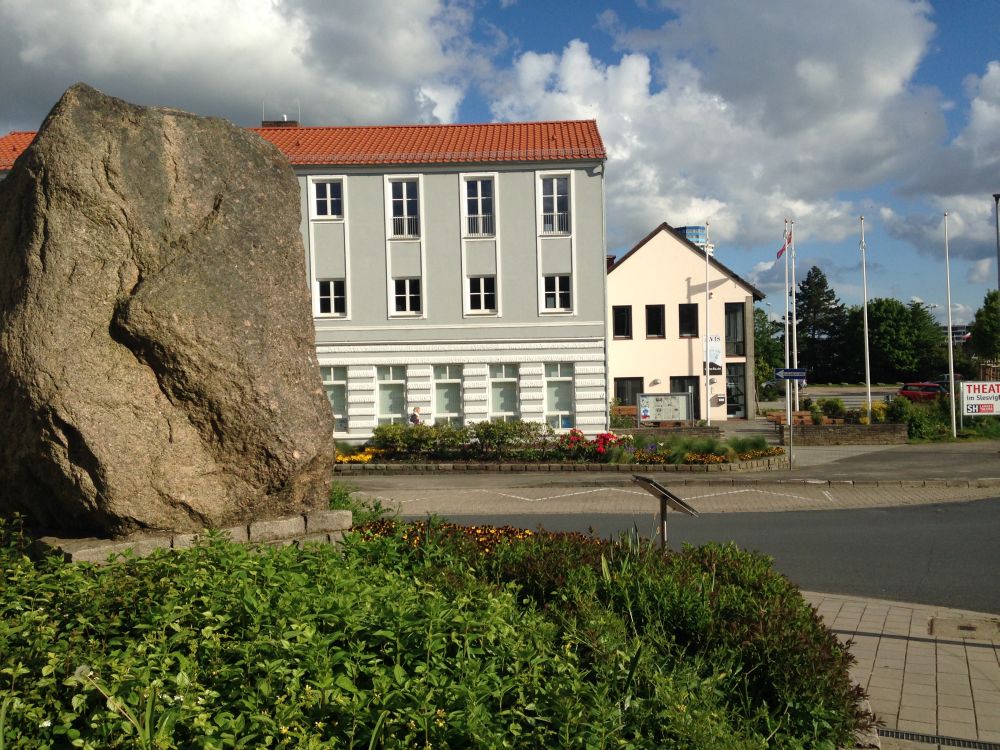 Im Vordergrund ist die Seite des Steins zu sehen, im Hintergrund das Slesvihus auf der anderen Straßenseite.